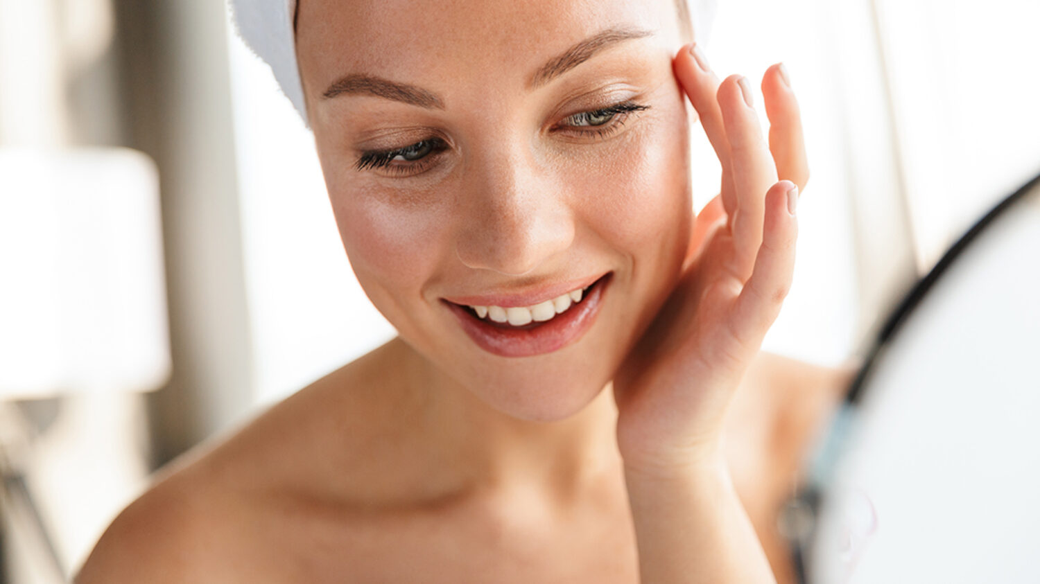 Photo of young joyful woman wrapped in white towels doing makeup while looking in mirror after shower